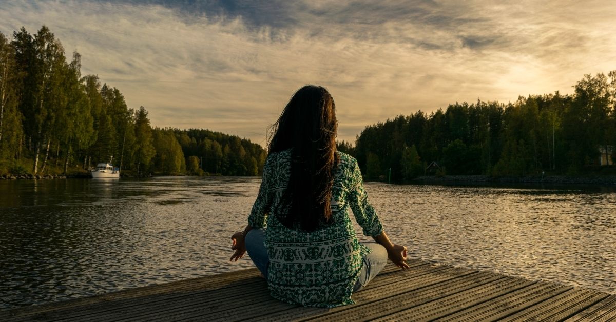 A woman meditating next to a lake and forest